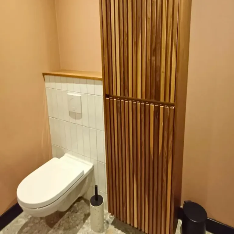 Een badkamer of toiletruimte met een wit hangtoilet en een hoge, slanke houten kast met verticale latten, passend bij een bijpassend badmeubel.