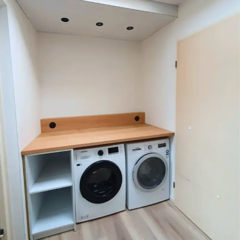 Een witte wasruimte met twee wasmachines onder een L-vormig houten werkblad, met daaronder witte open schappen en ingebouwde spots in het plafond.